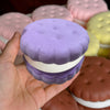 A hand holds a purple cake squishy fidget toy with white frosting, surrounded by other pastel-colored squishies.