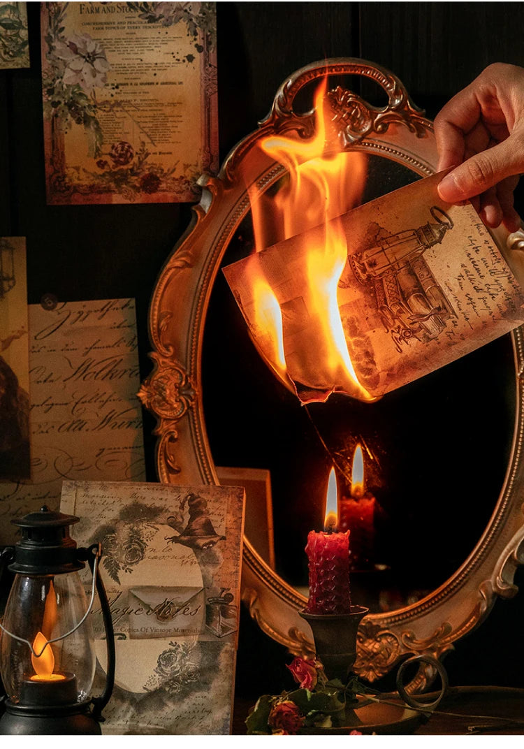 A hand holds an antique-style mirror reflecting a burning piece of aged paper and a lit candle.