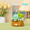 A small wooden flower basket made of building blocks holds miniature pastel-colored flowers and greenery.