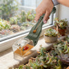 A hand holds a green handheld insect catcher with a magnifying lens, positioned near potted succulents by a window.