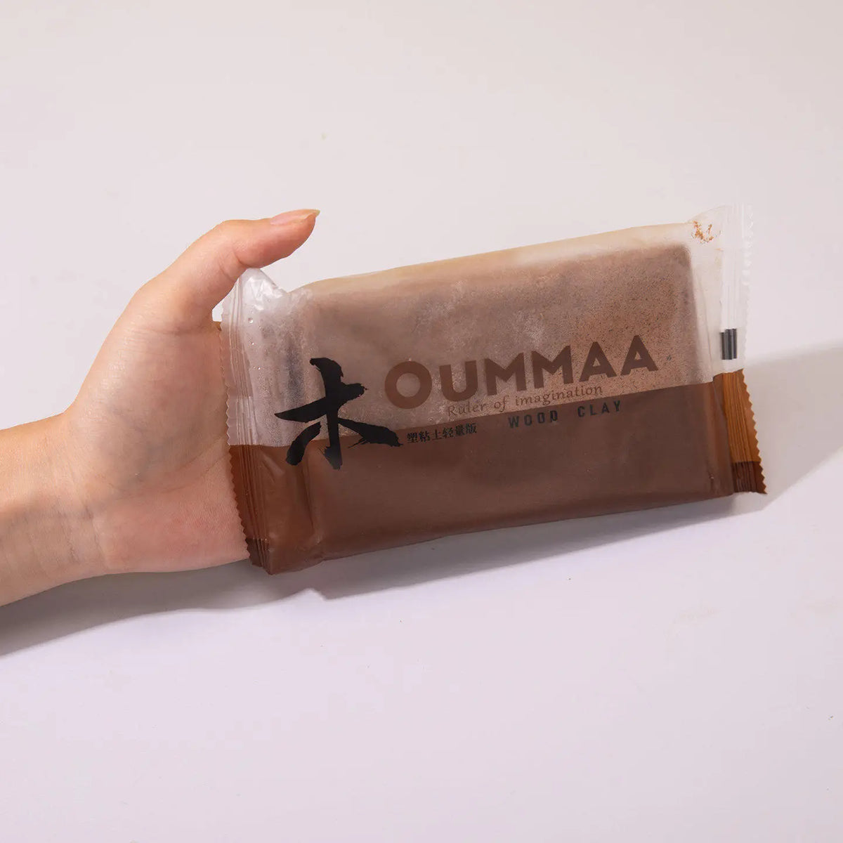 A hand holds a small package of OUMMAA wood clay for sculpting.