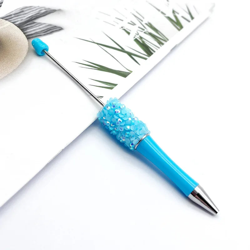 A close-up of a bright blue ballpoint pen with a cluster of blue sequins on the tip, resting on white paper.