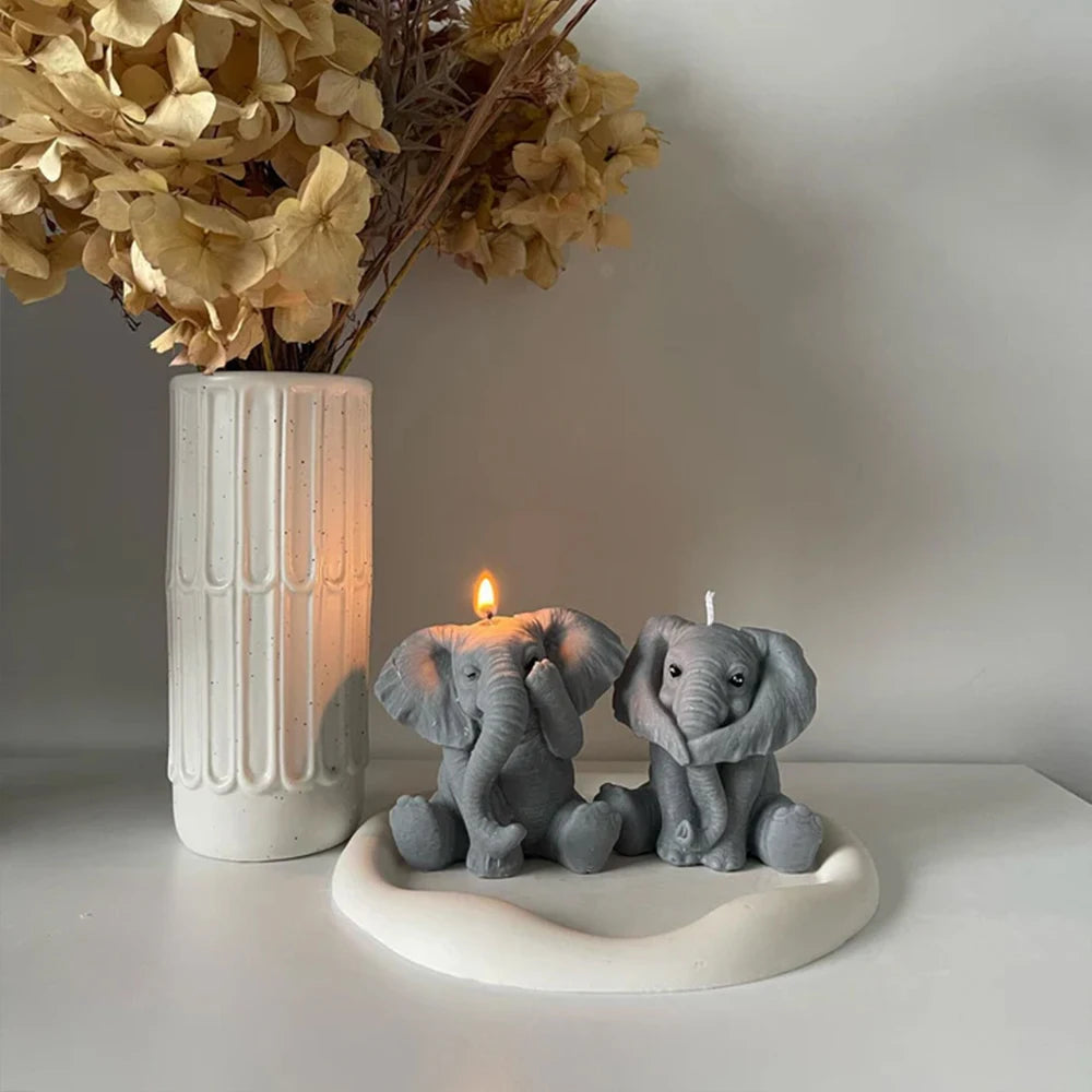 A gray silicone elephant mold sits on a white dish next to a lit candle and dried floral arrangement.