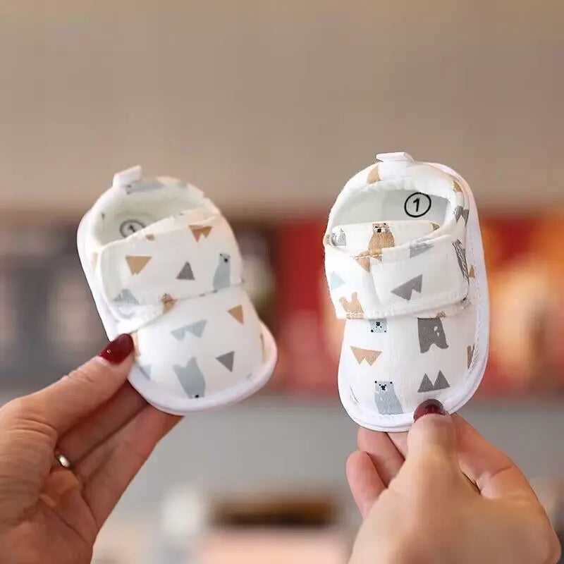 A pair of white soft sole first walker shoes for babies with bear patterns being held by a hand.