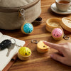 A hand holds a yellow dumpling squishy keychain with an LED glow on a wooden table next to a notebook and other accessories.
