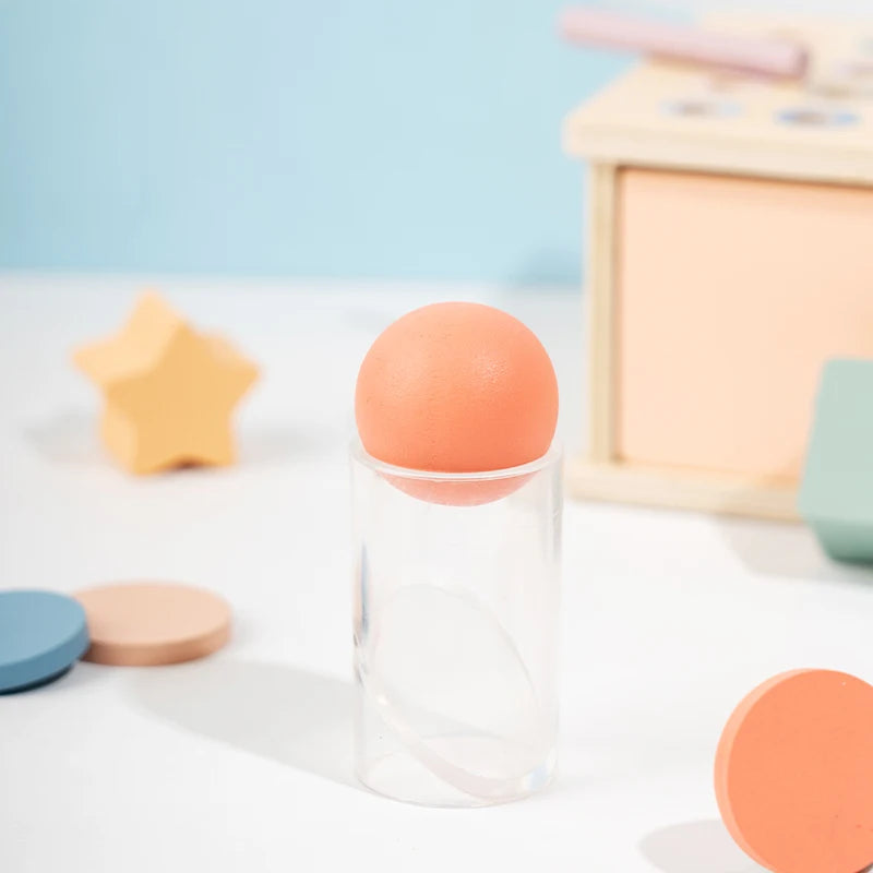 An orange egg is displayed in a clear glass jar on a white surface next to a wooden Montessori sorting box.