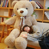 A young woman is resting her head on a giant brown plush teddy bear sitting on a light-colored sofa surrounded by books.