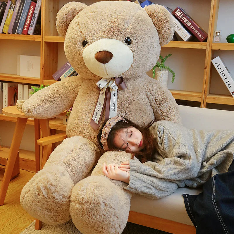 A young woman is resting her head on a giant brown plush teddy bear sitting on a light-colored sofa surrounded by books.