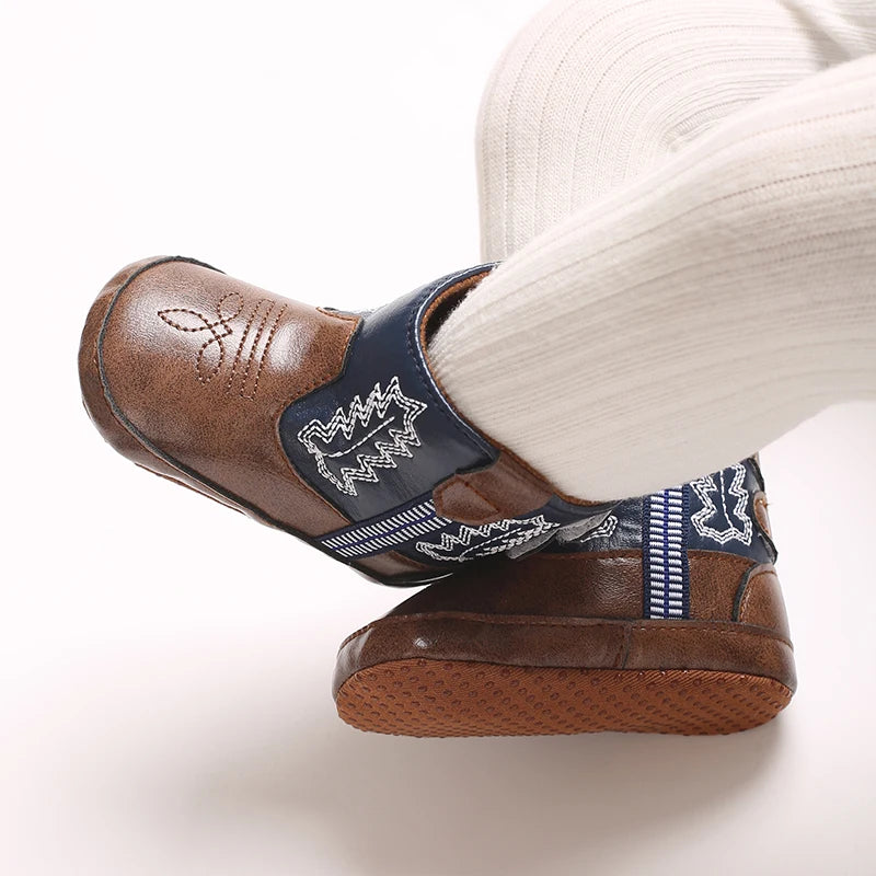 A close-up of a brown baby western bootie with a blue trim and soft non-slip sole worn on a person's foot.