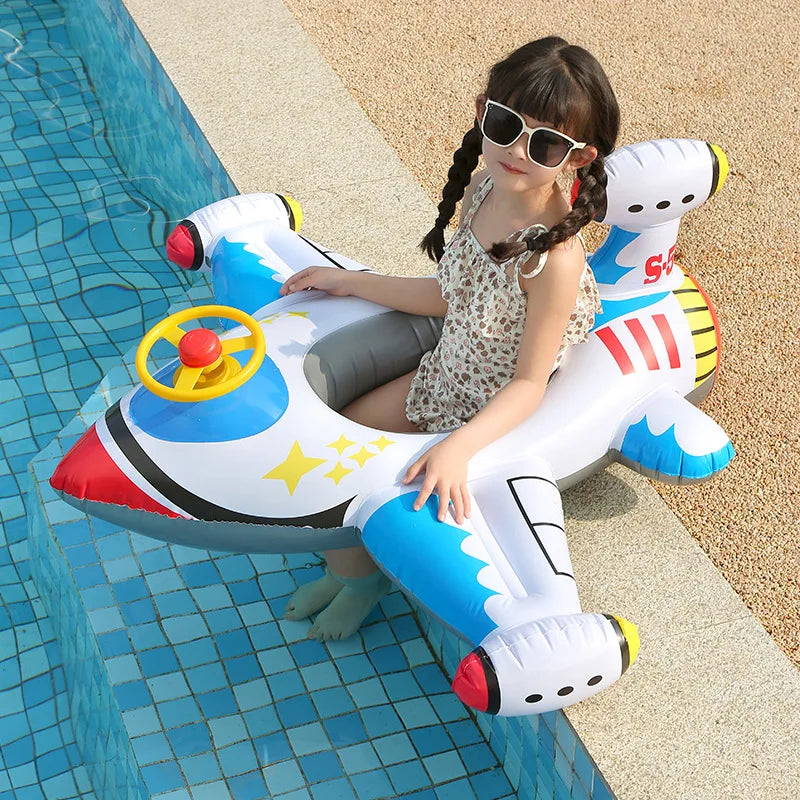 A young girl is sitting in a white and blue inflatable aircraft swim ring on a tiled pool deck.