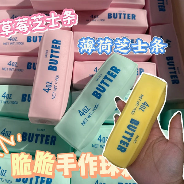 Eine Hand hält einen gelben Butter-Squishy-Stressball neben verschiedenen rosa und hellgrünen Butterstäbchen.