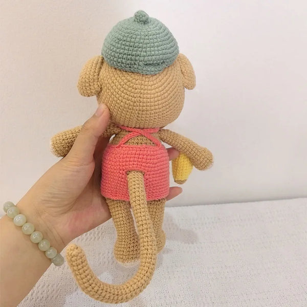 A handmade crochet monkey plush toy with a light blue cap and pink outfit being held by a hand.