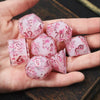 A hand holds a set of pink and white resin polyhedral dice showing numbers like 12, 70, and 6.