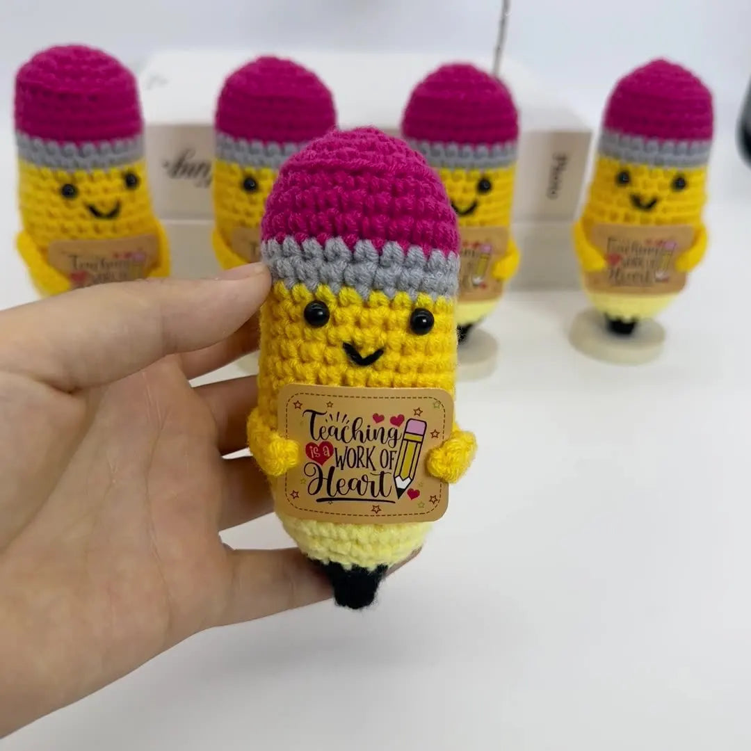 A hand holds a yellow crochet pencil ornament with pink and gray yarn details, displayed in a box.