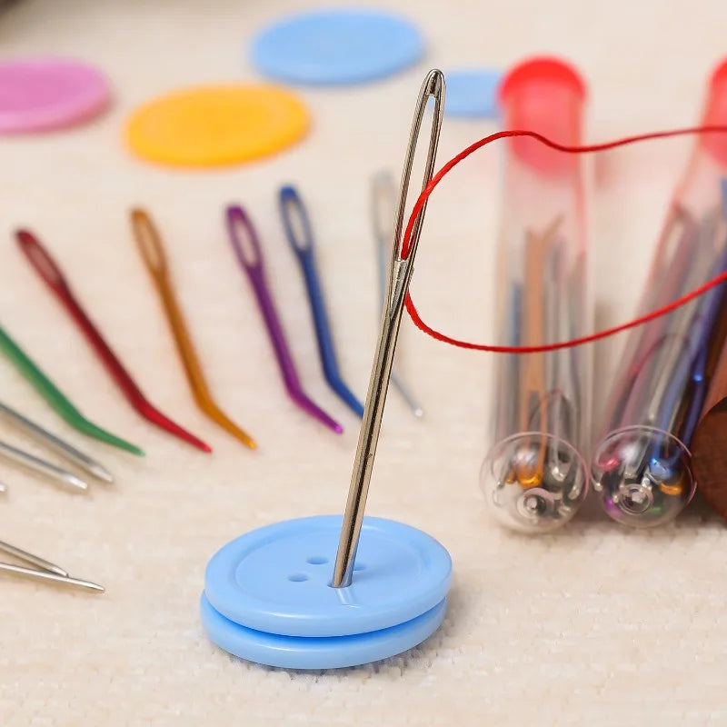 A stainless steel sewing needle is shown with a red thread, resting on a blue button, next to various colored needles and a s