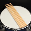 A set of light wood maple drumsticks laid diagonally across a white drum head.