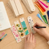 Hands are decorating blank wooden bookmarks with colorful tassels and paint on a light wooden surface.