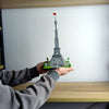 A hand holds a miniature model of the Eiffel Tower built from green building blocks against a white background.