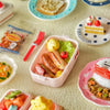 A collection of miniature food items and a doll picnic lunch box with a handle on a light surface.