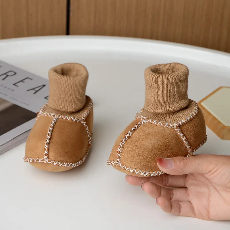 A pair of tan suede baby booties with a ribbed cuff and white stitching are shown resting on a white surface.
