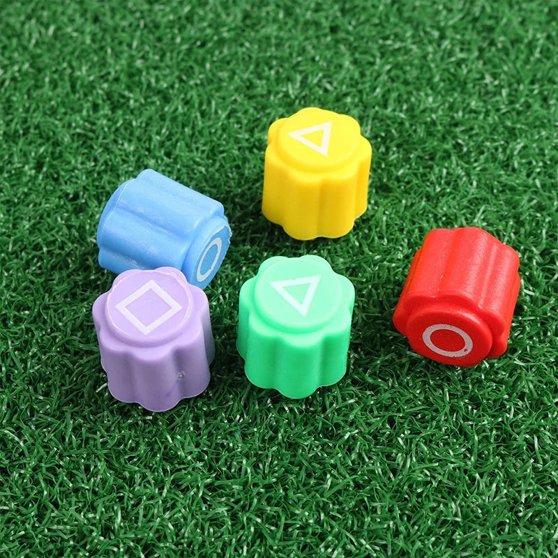 A set of five colored gonggi stones, including yellow, blue, purple, green, and red, resting on artificial green grass.