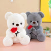 A white plush bear and a gray plush bear with red rose accents sitting on a pink surface.