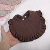 A hand holds a brown ruffled muslin cotton baby bib with an adjustable snap closure.