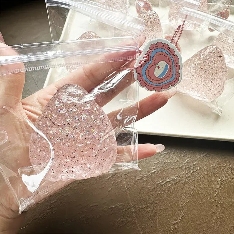 A hand holds a clear plastic bag containing a pink glitter strawberry squishy toy and a heart-shaped charm.