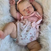 A newborn baby doll with hand-painted skin is sleeping on a fluffy white surface.