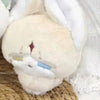 A soft, light beige and white collectible plush pendant doll with a visor detail resting on a woven surface.