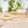 A wooden baby brush and comb set with the name Dhylicia engraved on the handle, resting on a light fabric.