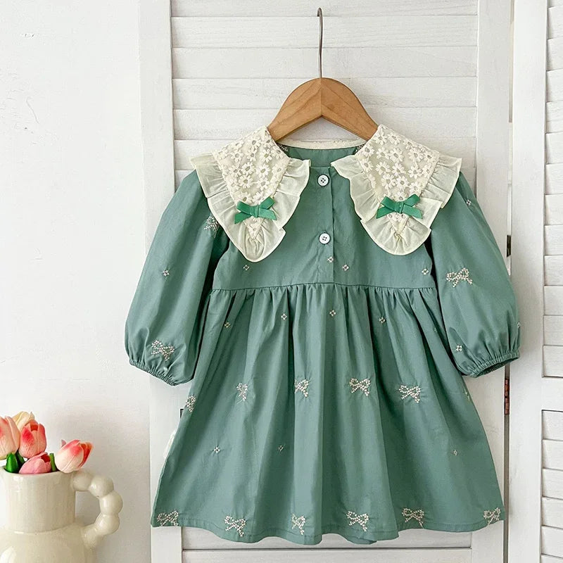 A sage green girls' party dress with white lace collar details hanging on a wooden hanger.