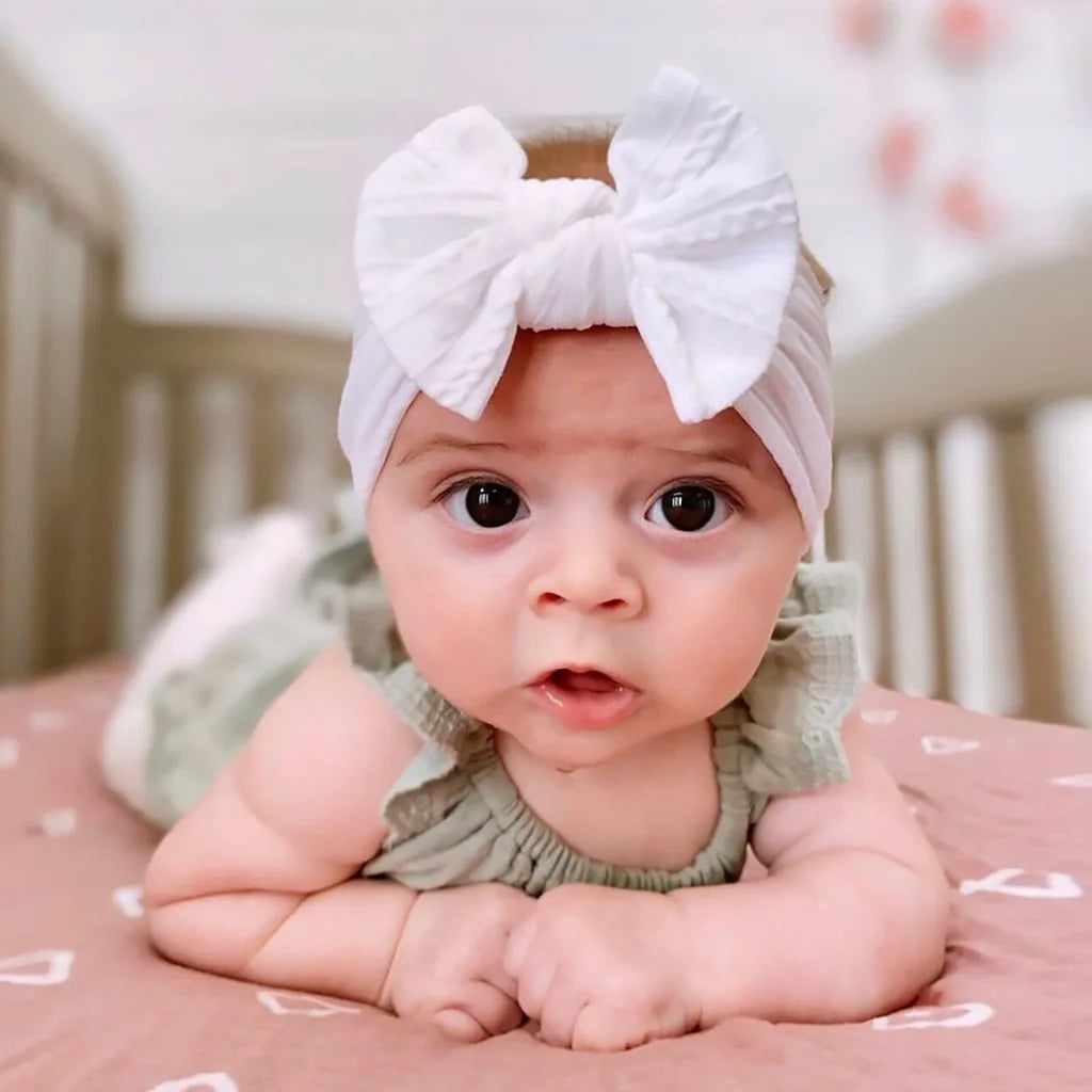 A baby wearing a white bow headband, looking directly at the camera.