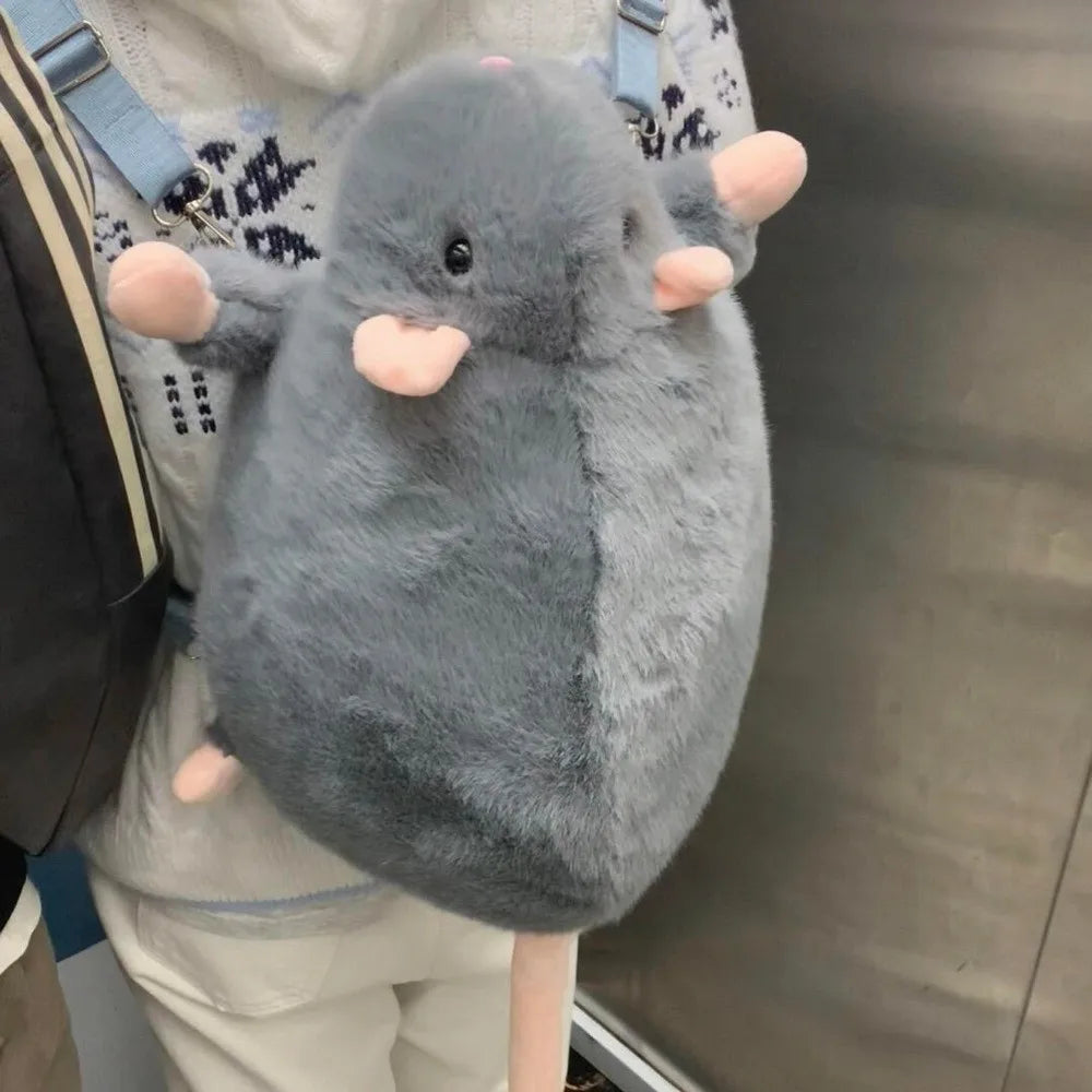 A gray mouse plush toy with pink ears is being held against a person's white shirt and backpack.