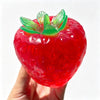 A bright red strawberry squishy stress ball with green leaves is held in a hand.