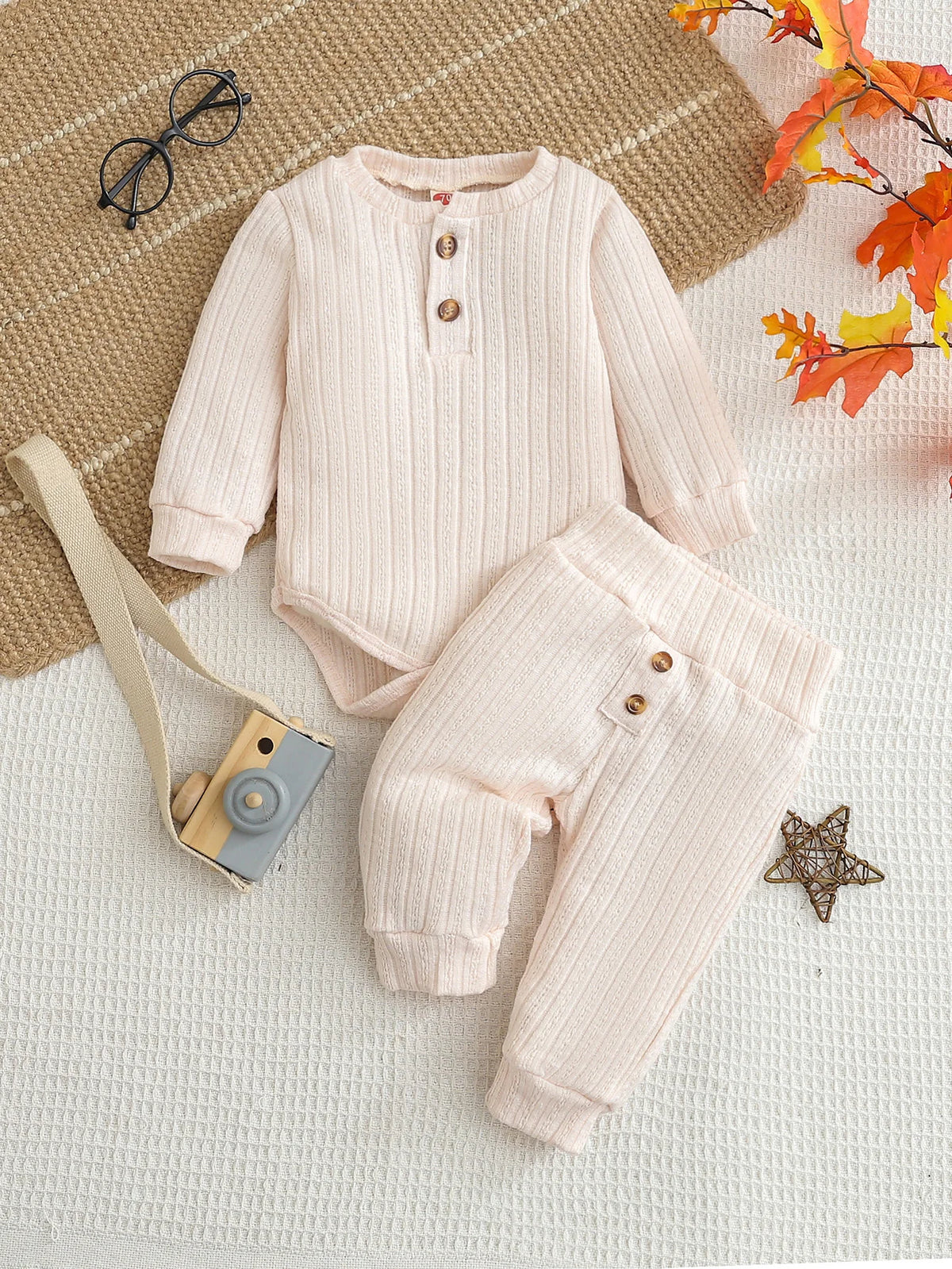 A light pink knitted baby romper and pants set laid on a white surface with natural fibers and autumn leaves.