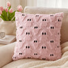 A pink fuzzy pillow with black ghost patterns rests on a white blanket next to a vase of pink tulips.