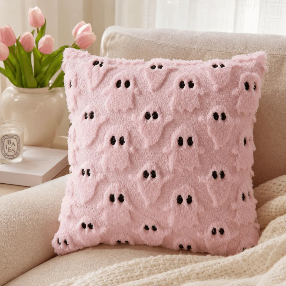 A pink fuzzy pillow with black ghost patterns rests on a white blanket next to a vase of pink tulips.