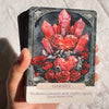 A hand holds a Garnet crystal oracle card featuring a red heart shape surrounded by roses and dark background.