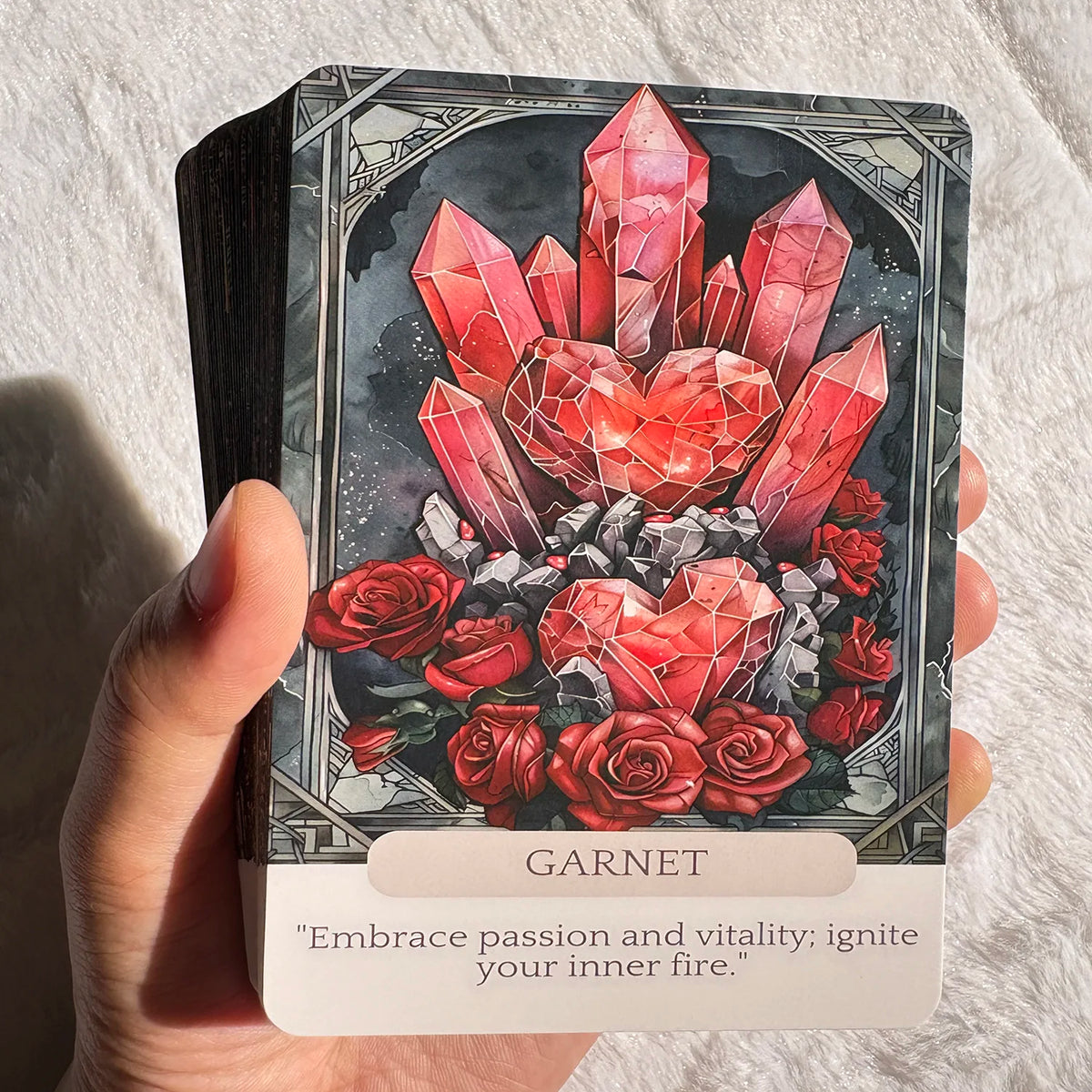 A hand holds a Garnet crystal oracle card featuring a red heart shape surrounded by roses and dark background.