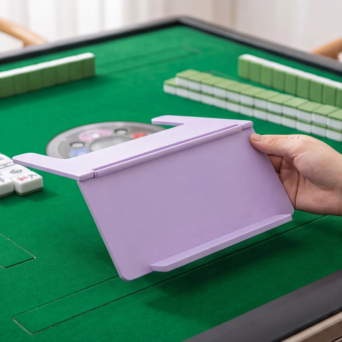 A hand holds a purple foldable mahjong card holder with a non-slip stand on a green table.
