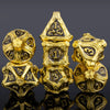 A set of seven gold-colored metal dice, including a skull and dragon design, displayed on a dark surface.