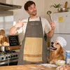 A man wearing a gray chef apron with a pocket is pointing, standing in a kitchen setting.