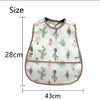 A white baby bib with a green cactus pattern and a brown trim, shown next to a measurement scale.
