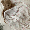 A soft, white cotton muslin swaddle blanket with a delicate pink floral print and a brown teddy bear toy.