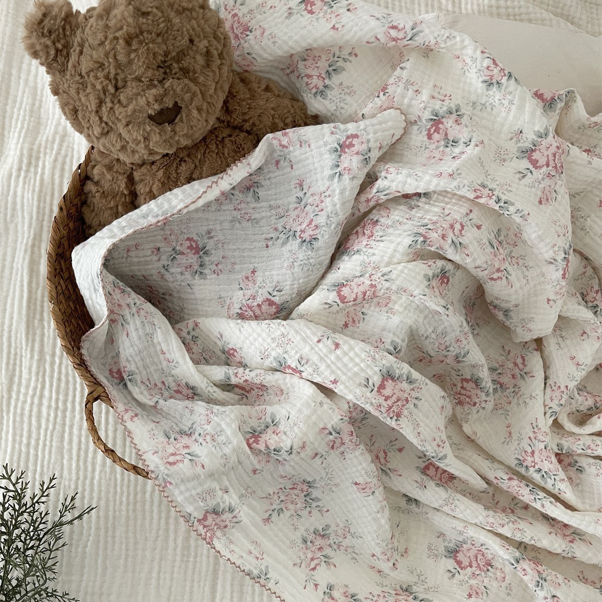 A soft, white cotton muslin swaddle blanket with a delicate pink floral print and a brown teddy bear toy.