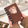 A brown sticker book with a dark trim and a small tag is held in a hand, set against a white background.