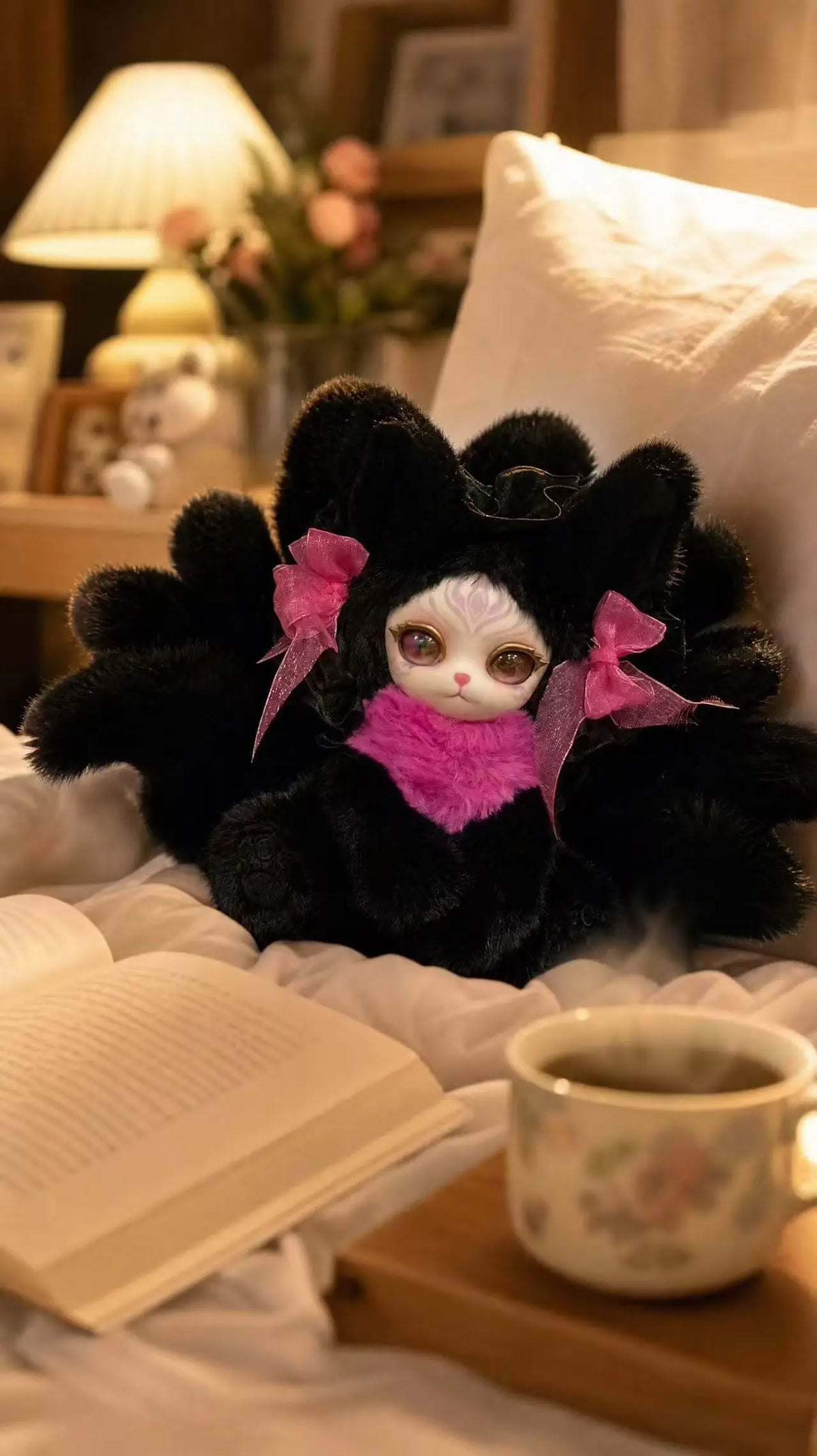 A black nine-tailed fox plush collectible with pink accents resting on white fabric next to an open book.