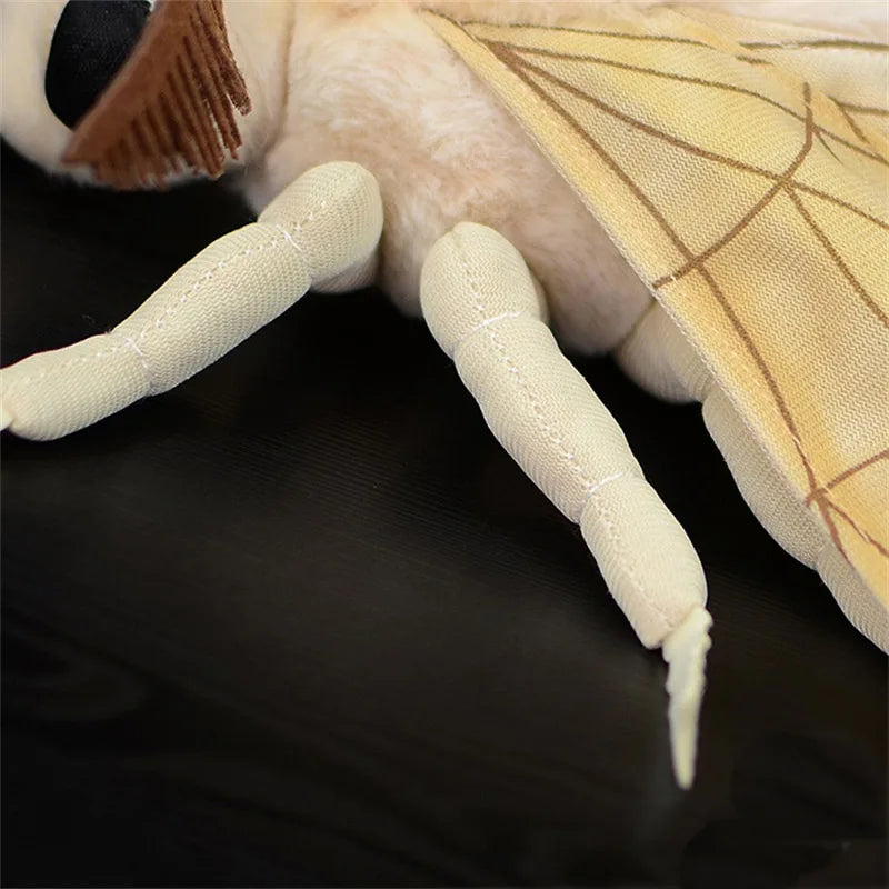 A close-up of a light beige silk moth plushie showing its soft body and patterned wing.