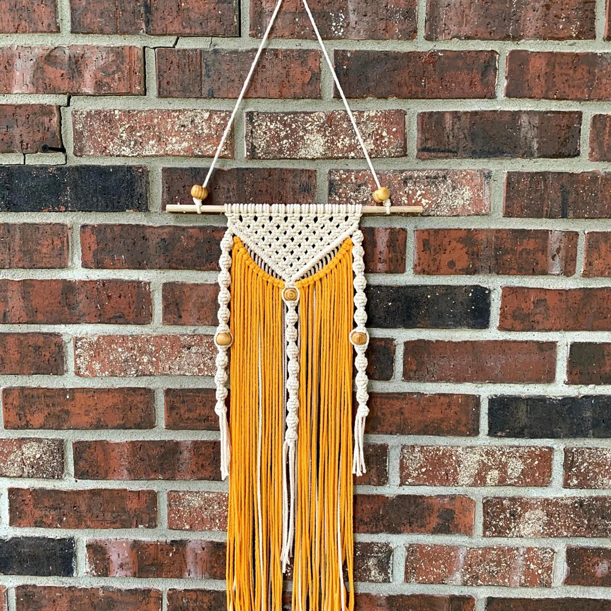 A hanging display of natural wooden spacer beads in yellow and white thread against a brick wall.